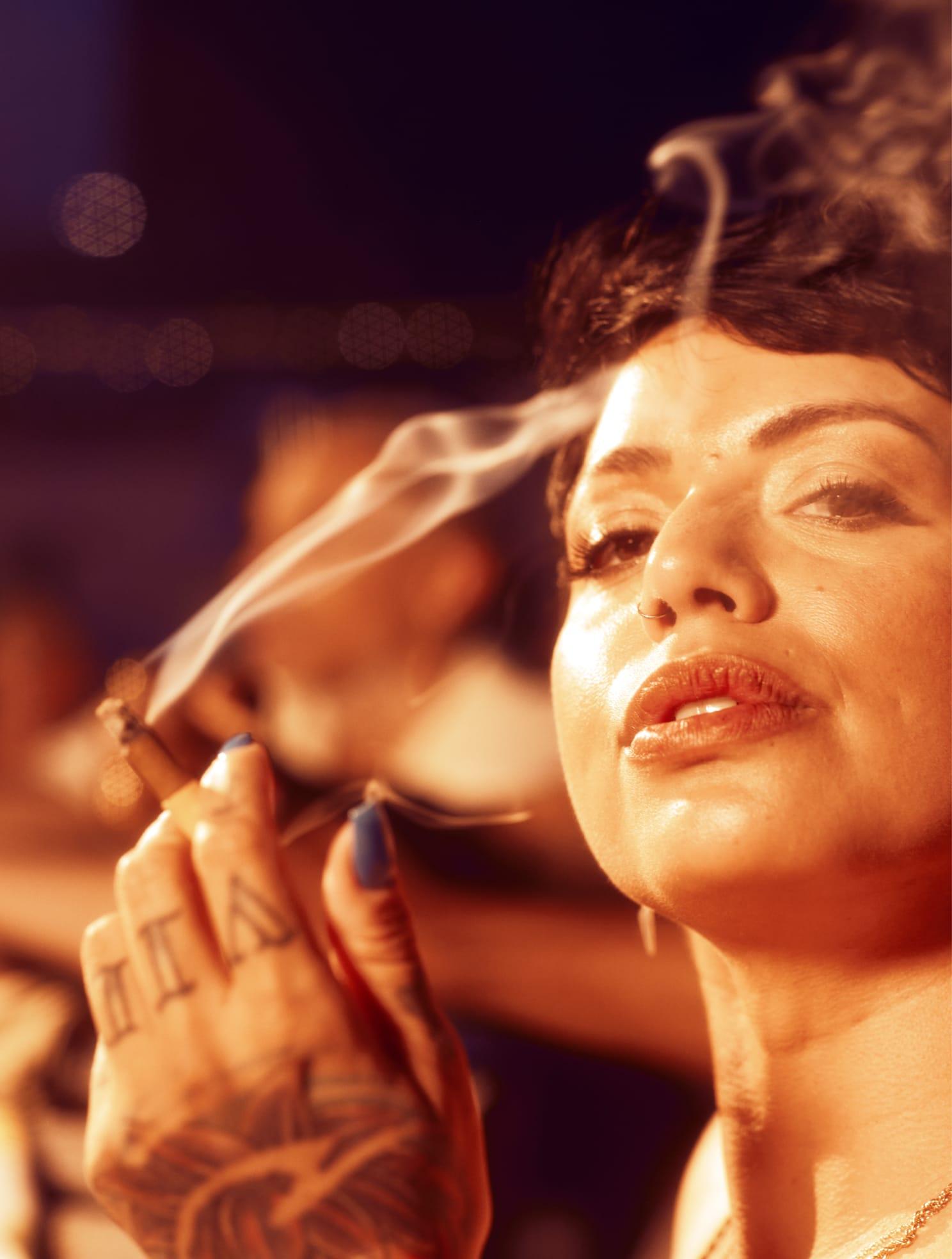 Close-up of a woman with short hair, holding a smoking blunt and gazing confidently, with soft lighting and a relaxed atmosphere.